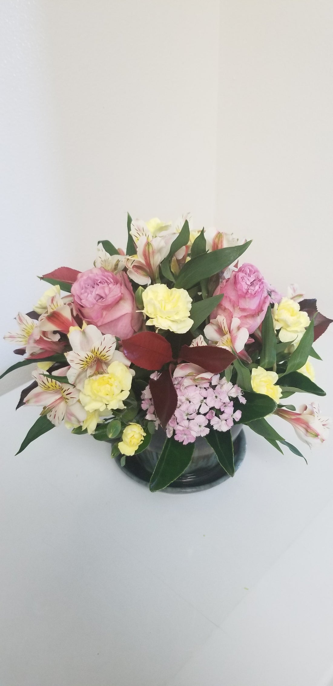 Fresh flower arrangement arranged in a tea cup. Queen's Crown garden roses present a lovely purple tone complementing yellow mini carnations, pink dianthus, pink alstromeria and lovely mixed foliage.