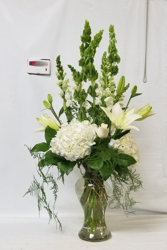 A captivating white and green floral arrangement featuring an exquisite blend of roses, hydrangea, lilies, snapdragons, Bells of Ireland, and flowing greens, creating a harmonious and elegant display